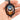 A hand holding an oval pendant necklace with a black ornate frame and a colorful stained-glass-style design featuring a bat silhouette in blue, purple, orange, and yellow.