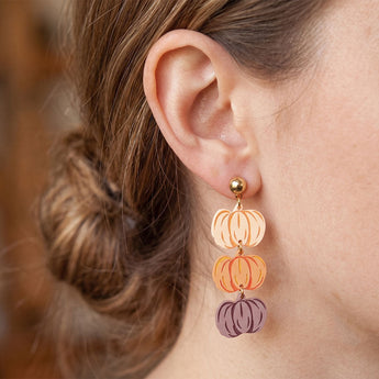 A close-up of a person's ear wearing a dangling earring with three hand-painted pumpkin charms in peach, orange, and muted purple, suspended from gold-toned metal.