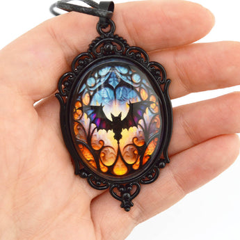 A hand holding an oval pendant necklace with a black ornate frame and a colorful stained-glass-style design featuring a bat silhouette in blue, purple, orange, and yellow.