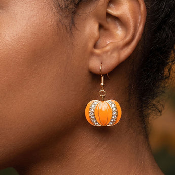 Close-up of a person's ear wearing a dangling pumpkin-shaped earring in bright orange with clear rhinestone trim and a gold hook.