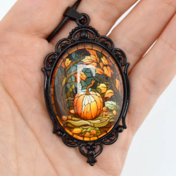 A hand holding an oval pendant necklace with a black ornate frame and a stained-glass-style autumn scene featuring a pumpkin, leaves, and a white letter 'O' in the center.
