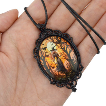 A gothic-style oval pendant necklace with a black ornate metal frame and a dark cord, held in a person's hand. The pendant features a colorful printed illustration of a ghostly figure in a spooky forest at night under a full moon.
